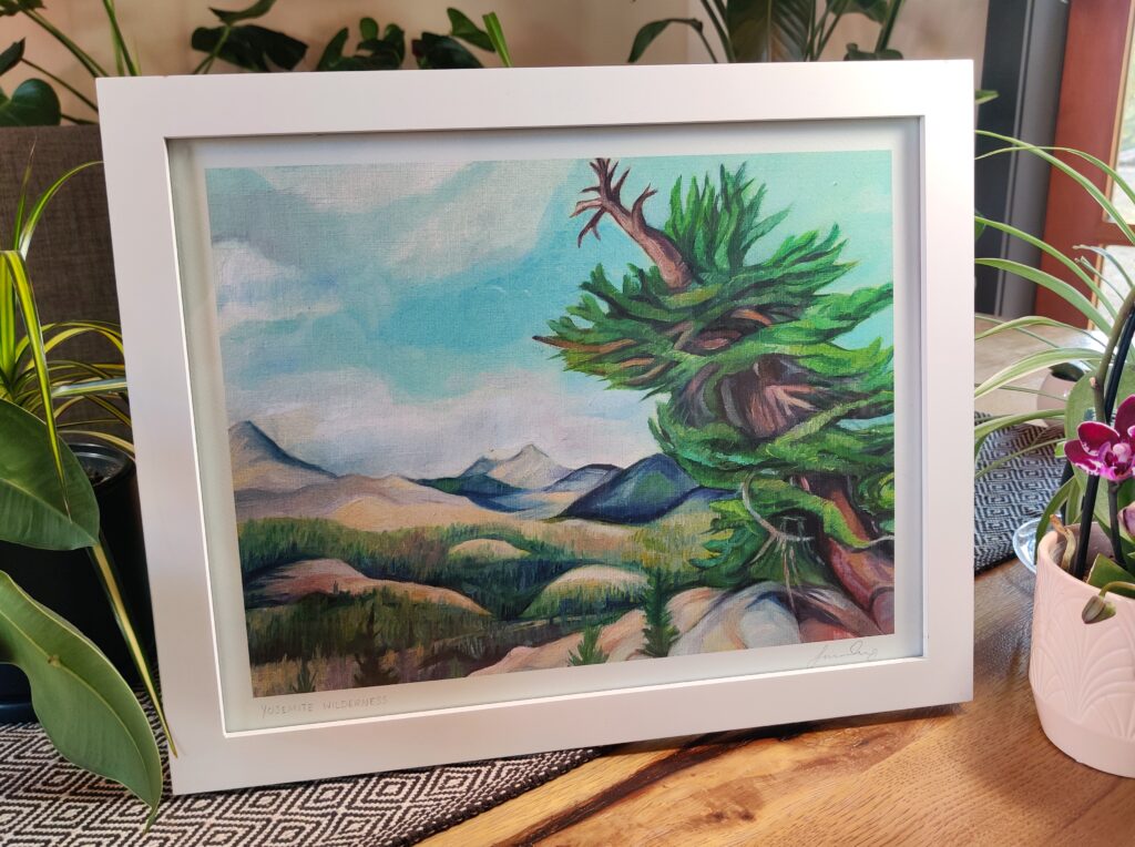 Painting of gnarled tree in Yosemite backcountry with mountains in the background, in frame on table.